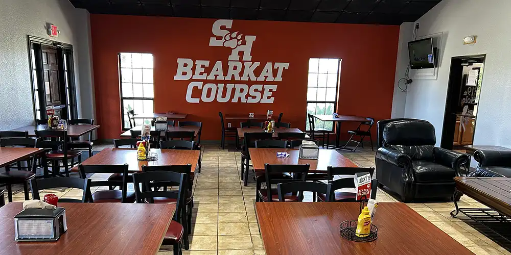 Interior of the SHSU Bearkat Course clubhouse dining area featuring tables and chairs, lounge seating, and a large orange wall displaying the Bearkat Course logo.